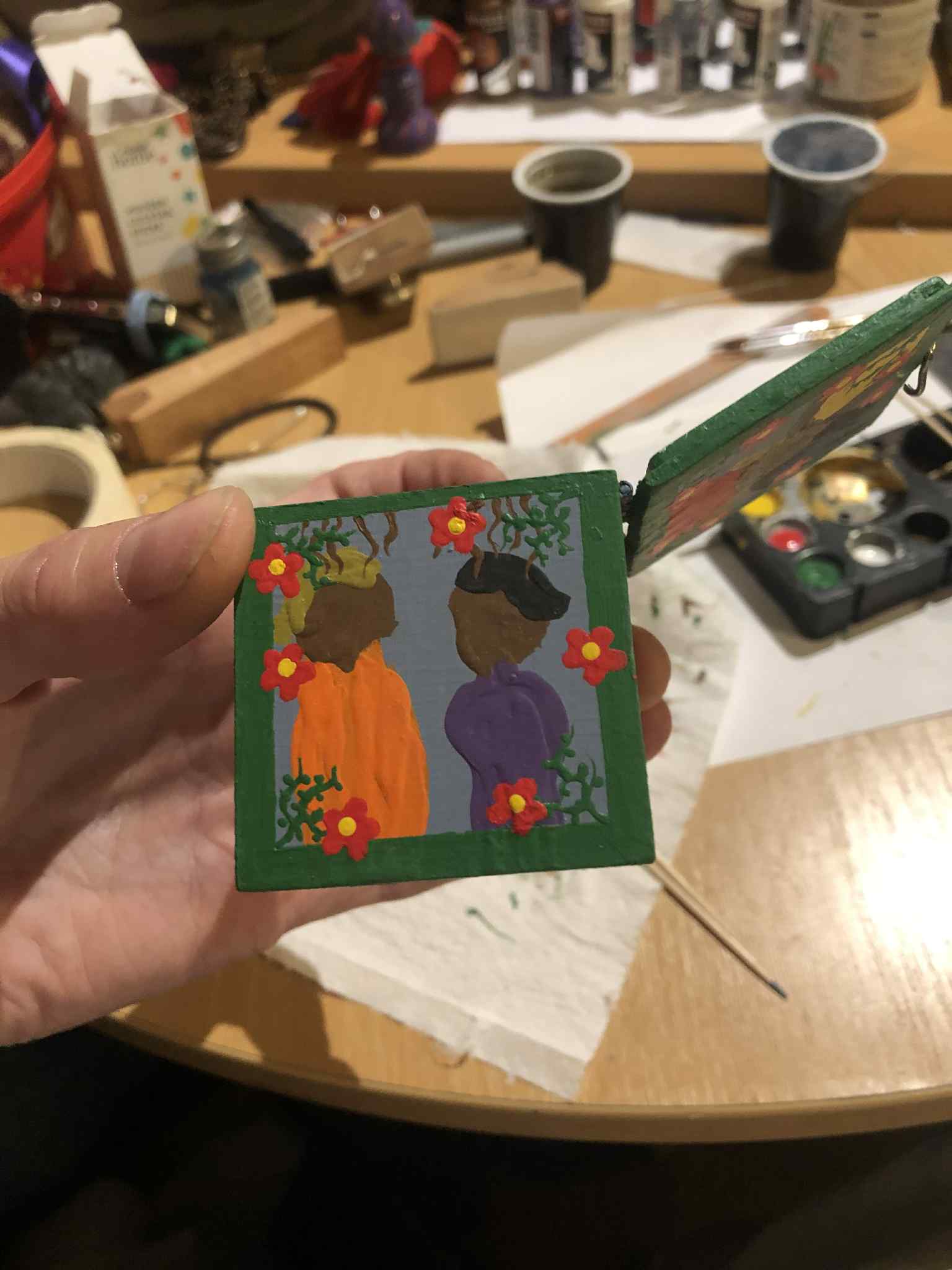 The same side as the last photo, with a figure wearing orange with blonde hair and a figure wearing purple with dark brunette hair, with brown tentacle fingers coming down towards and onto their heads from above, and the vinework in the corners and red flowers with yellow centers spread out around the green borders of the side.