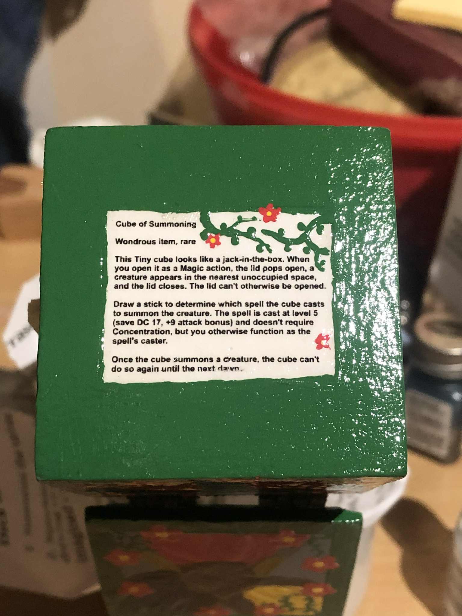 The bottom of the box, glossy with its first coat of varnish, showing the instructions in tiny text in the center of the face, which is painted fully green otherwise, being the bottom, with small green vinework added into the empty area of the printout beside the cube of summoning title, with three red flowers with the yellow center added as well.
