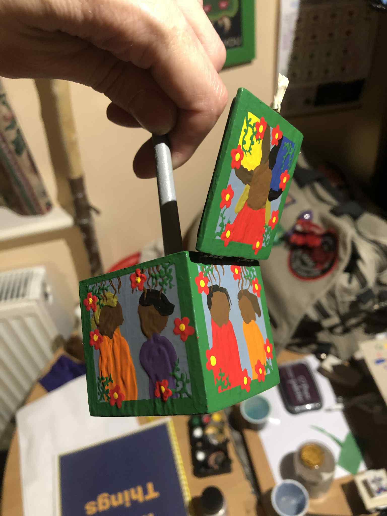 The box, covered with half of the layers of varnish that it will be getting, being held up by a grey stick, actually a paintbrush now covered in primer, which is taped to the inside of the box. The sides are glossy and still textured with the impasto, showing three sides due to the lid being opened, orange and purple wearing figures on the left, red and orange wearing figures on the right, and the three figures of red, yellow, and blue clothing with the one head on the top. Kabutroid's messy worktable is seen beneath.