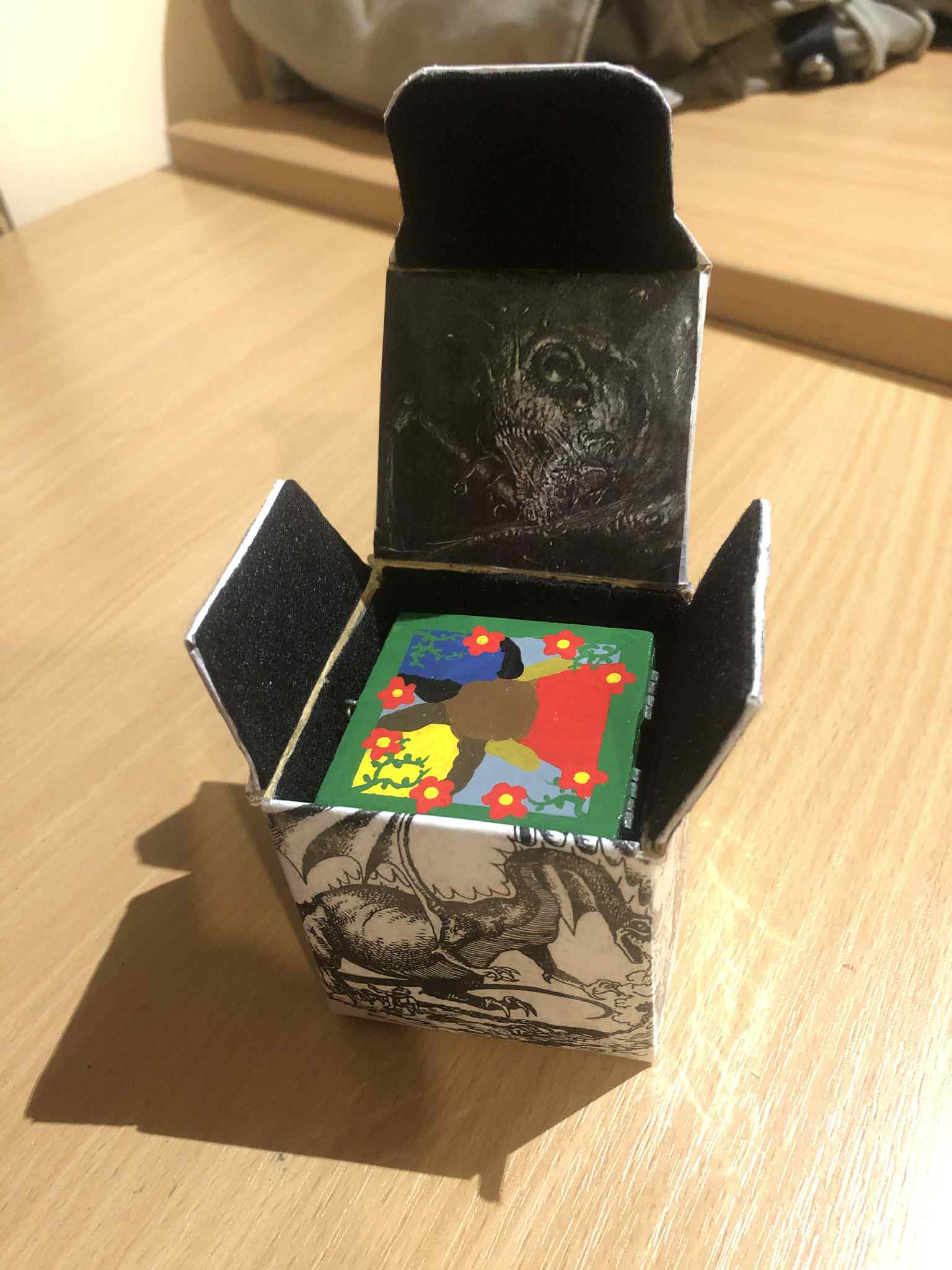 The inside of the cardboard box, showing a scary spider creature on the underside of the lid, and the inner flaps and sides all coated with a black felt fabric, and inside is the cube of summoning, brightly painted in its greens and reds inside the scary dark box.