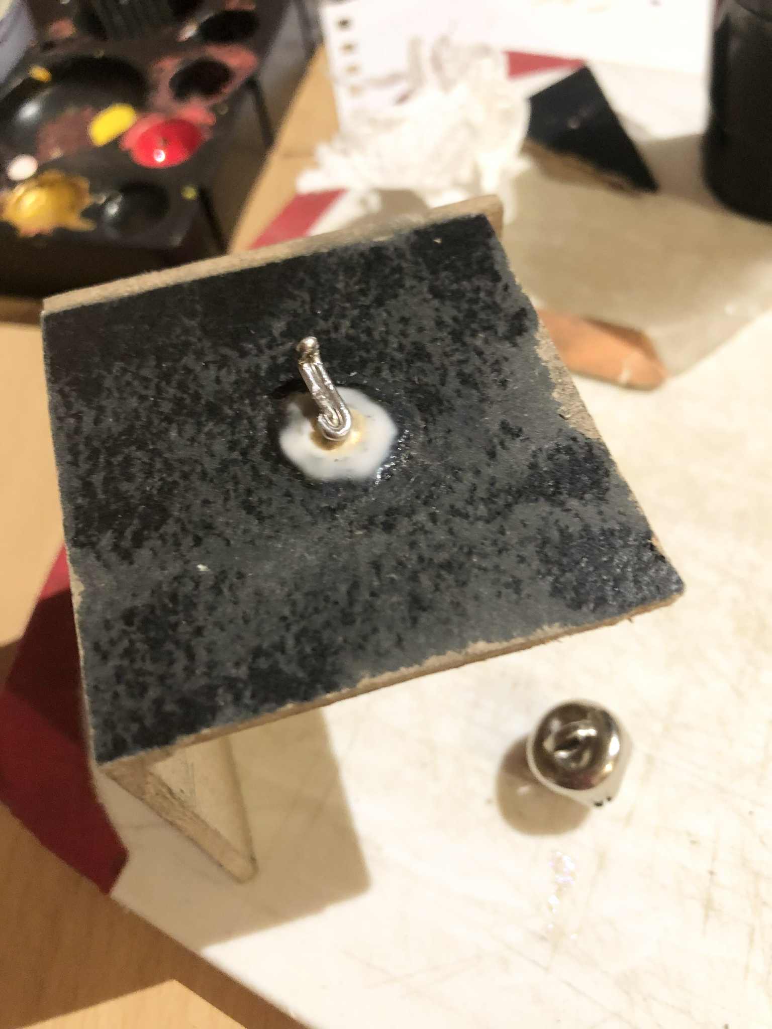 The crank is installed through the side of the box, a bent piece of wire which is also doubled for the crank portion itself, with the support washer being glued to the box, and the jingle bell on the work table below.