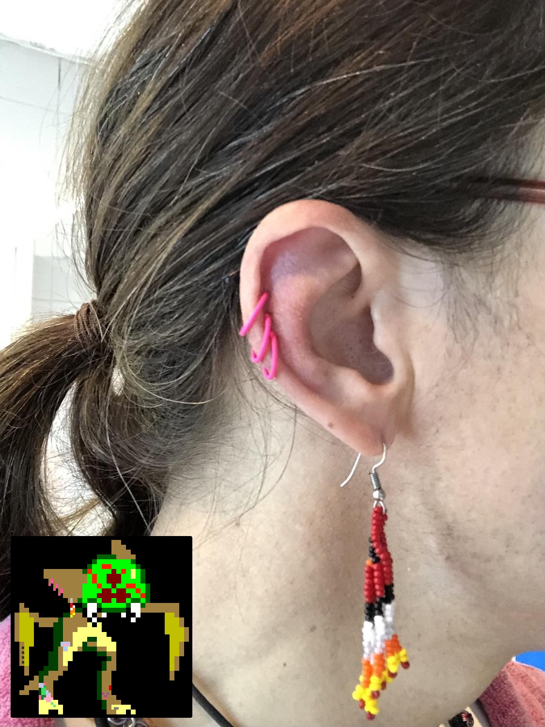 A photo of Kabutroid's right ear showing the three pink hoop rings in her helix piercings, while also wearing an indigenous red beaded earring in the lobe. In the bottom left corner, superimposed is Kabutroid's sprite, also showing a sprite version of the three hoops and beaded earring.