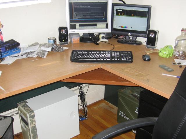 The computer setup, a brown wooden corner desk with two monitors, some general scatter on the desk, and the computer with a taped up faceplate is sitting under the desk.