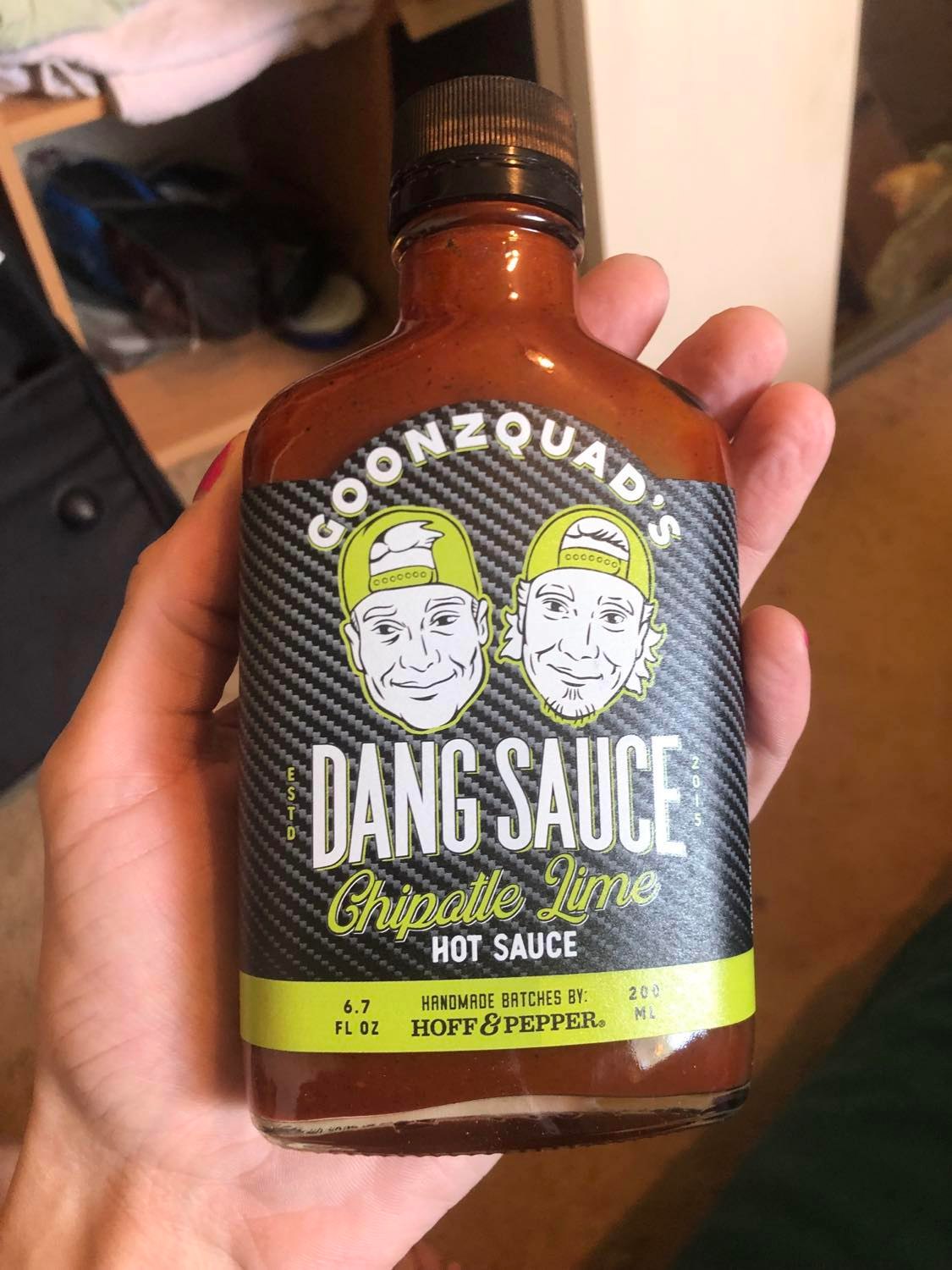 The front of the bottle, carbon-fiber backdrop, with caractures of the two goonzquad boys on top of it, their hats backwards. It's simple white with black linework, and the hats and outline are lime green. It boldly states DANG SAUCE, with Goonzquad's up at the top. Chipotle lime appear in script beneath dang sauce, and it is handmade batches by Hoff and Pepper. Six point seven fluid ounces.