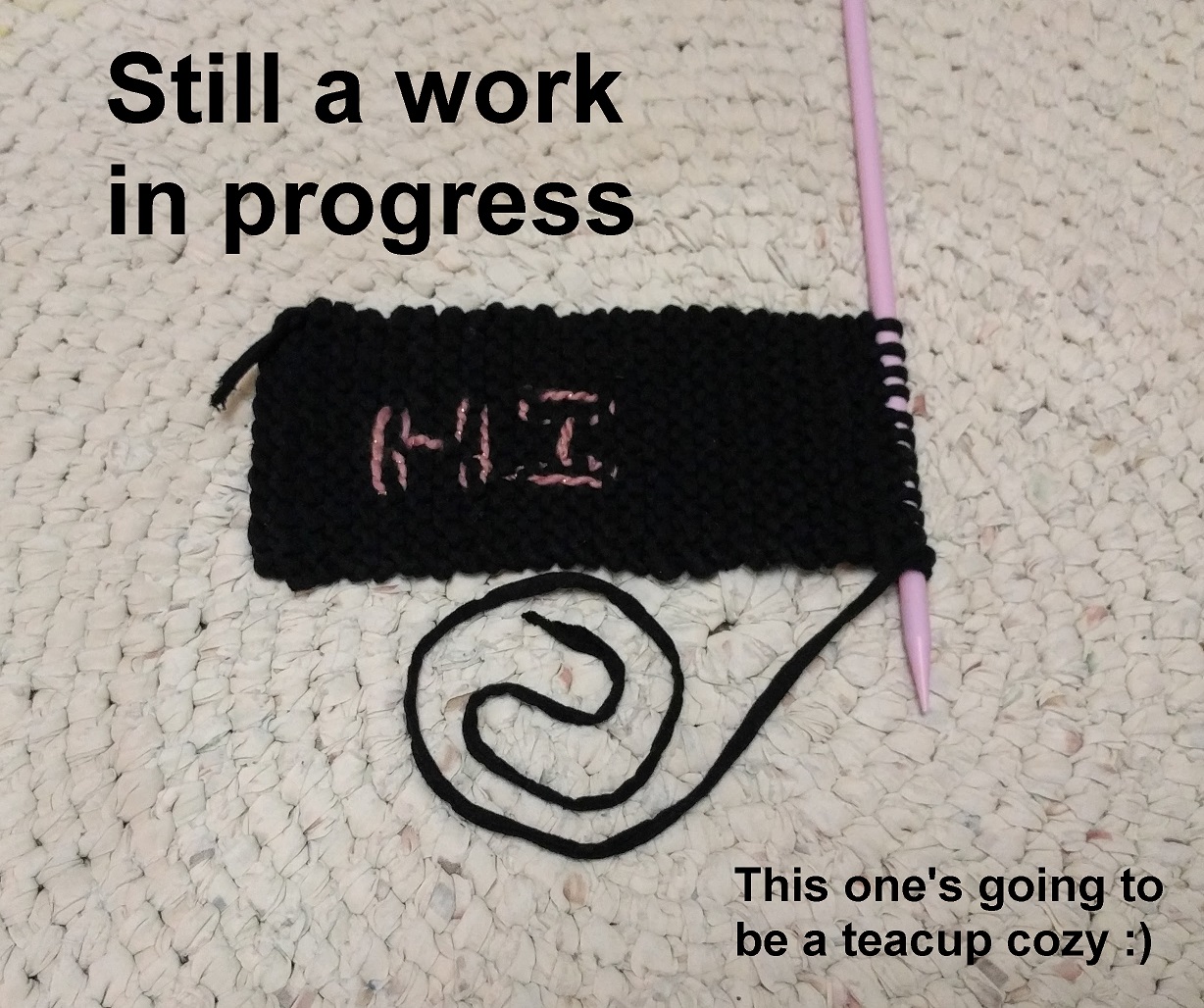 A small knitted rectangle with the word hi lightly sewn into it with pink yarn, and text saying still a work in progress.
