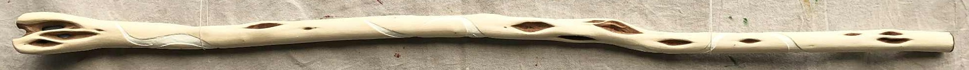 A peeled wooden staff with white linework running gently along the length of it. A large number of diamond patterns in the wood are prominent, and a light blue stone is embedded in the head of the staff, visible through three openings in the wood patterning.