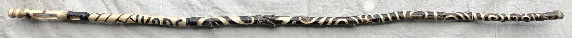 A dark brown wooden staff, with the majority of the bark still attached, with heavily swirling lines running along the length of it. The grip is made up from two distinct sections, one saying KABS, the other a brickwork pattern, and the head has a hollowed out mouth with a large staring eye above, and two curved sections above the eye.