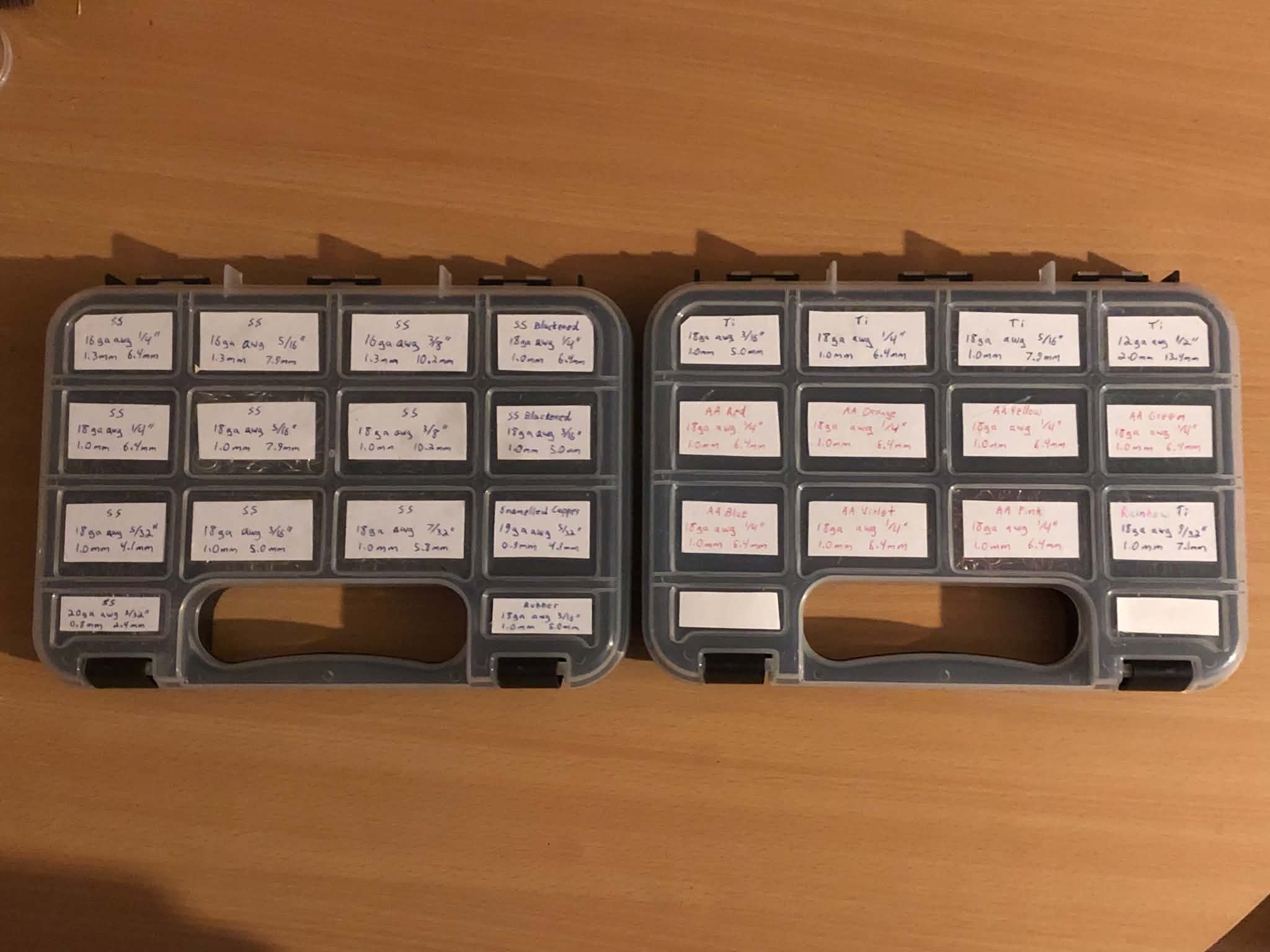 On a wooden table, two smaller cases with fourteen pockets each, labelled with blue, black, or red ink to indicate stainless steel, titanium, or aluminium rings, as well as their sizes in american wire gauge and metric sizes.