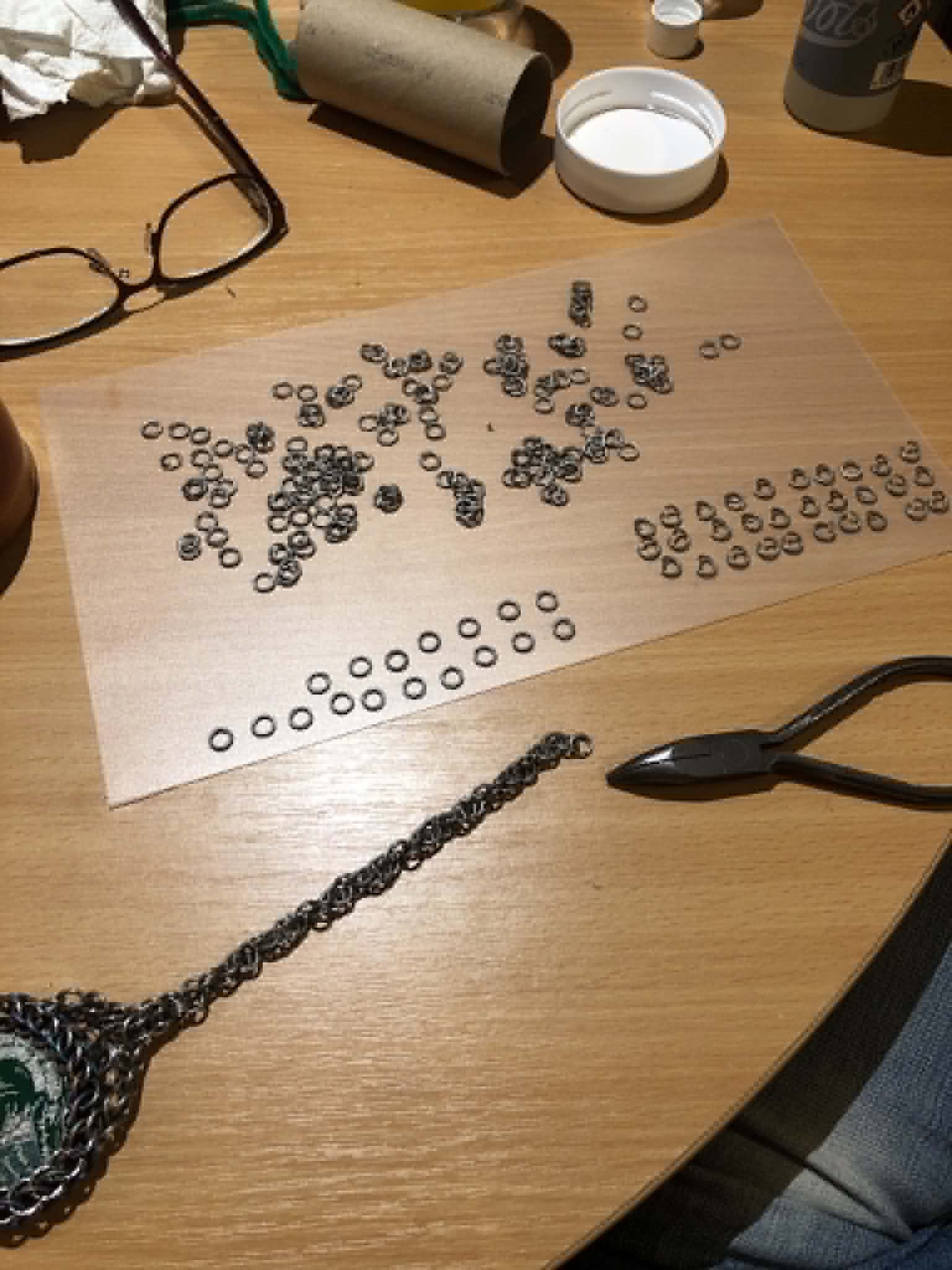Kabutroid's workbench, with all of the loose rings on a plastic sheet to dry after being rinsed from the first tumbling, many of them now pre opened or pre closed, and about three quarters of a foot of magician's chain now built and attached to the coin wrap. Also seen on the table is the full toilet roll tube that became the doohickey, Kabutroid's glasses, and a pair of needlenose pliers.