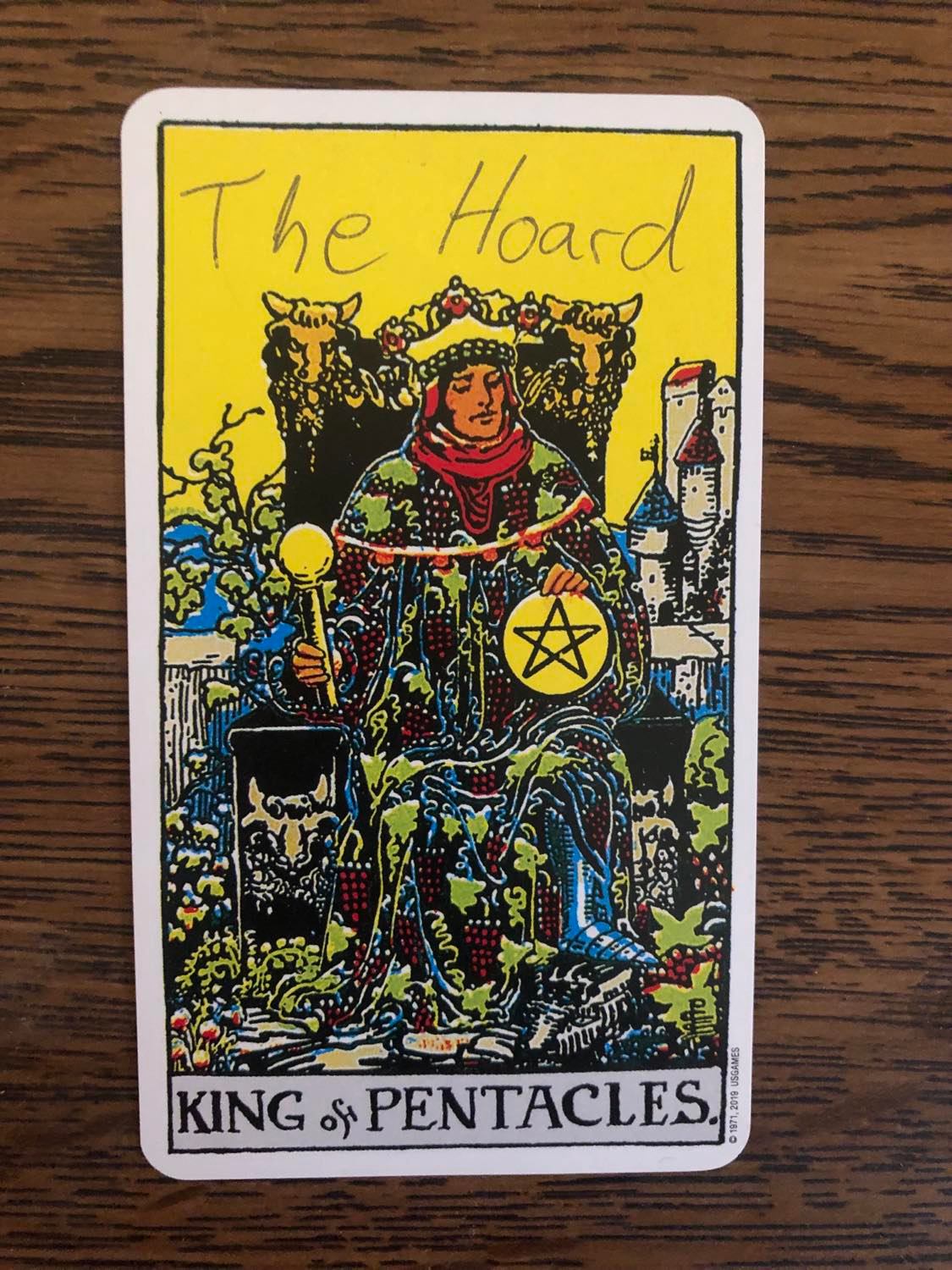 The king of pentacles card with the hoard written on it, laying on a wooden desk.