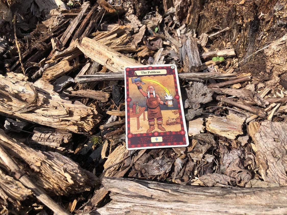 A man pouring a drink, the card laying on the forest debris.