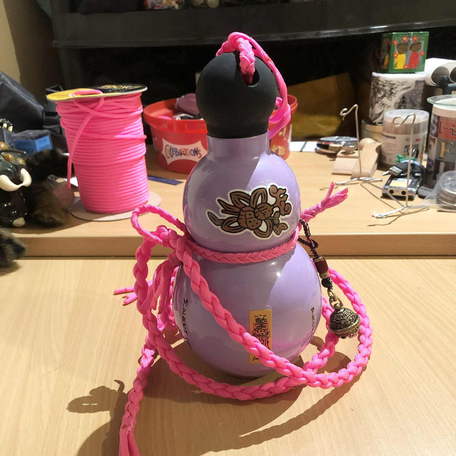A light pink purple lavender coloured, gourd shaped bottle, with a stylistic gourd knot on the top orb of the gourd, and the bottom has stickers with Japanese kanji for, as best Kabutroid can research, spiritual protection and balance, which came with the bottle. A small bell hangs on its side, and pink cord has been used to hold the cap and around the center, with a longer braided pink sas