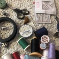 A white crocheted rug with on it a pile of different colour spools of thread, a pack of different sizes of needles, several sets of household repair needles, a spool of fishing line, scissors, a piece of leather, a measuring tape, and bobbins and a footer for a sewing machine.