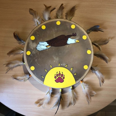 A tanned leather hand drum with fringe feathers around the rim, the face painted with an eagle flying overtop of a sun with a burgundy bear paw on it, and surrounded by smaller suns.