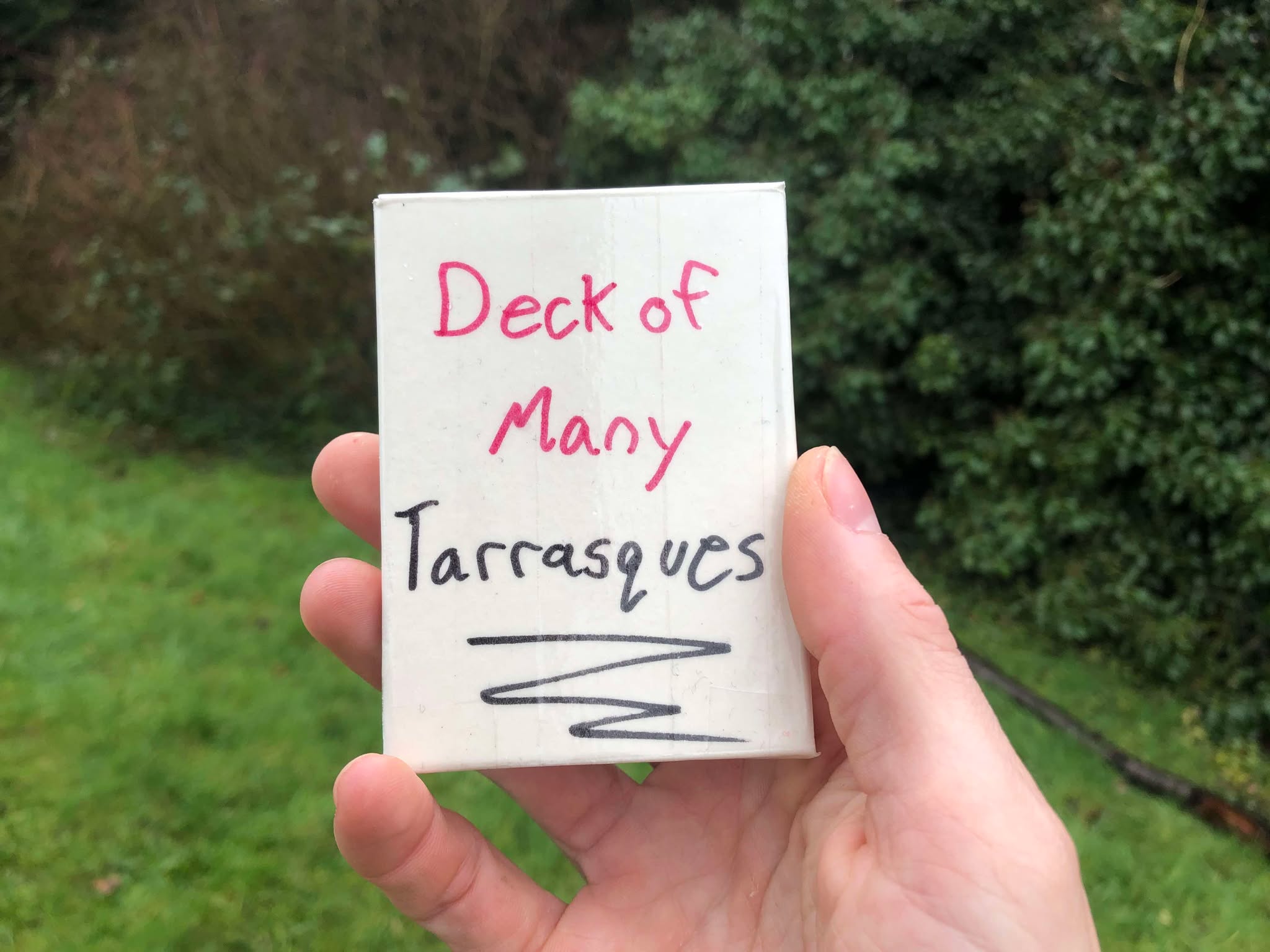 A deck of cards being held up in a grassy patch of a forested area. A Deck of Many Tarrasques. What might be inside, what could happen?