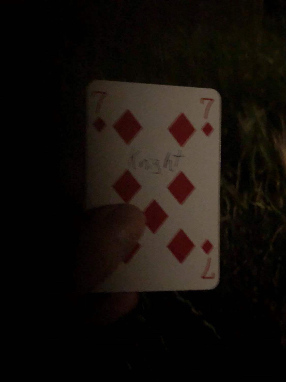 The seven of diamonds with the word Knight written on it, held in the barely lit dark.