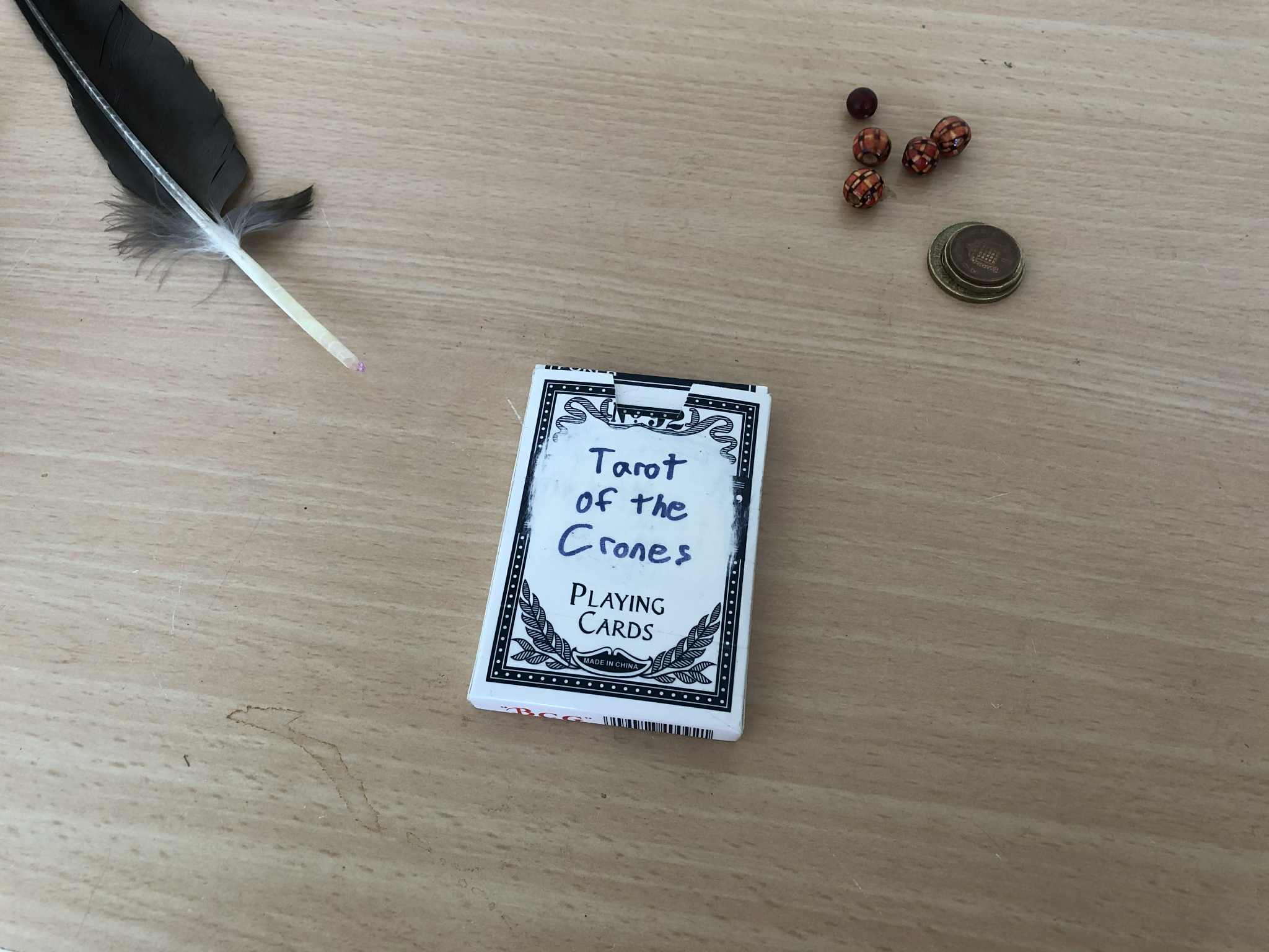 A deck of cards, the Tarot of the Crones, on a merchant's table beside a feather quill, a handful of coins, and several coloured gemstones.